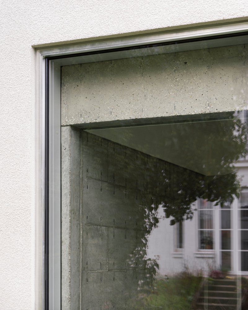 Window detail showing concrete lintel meeting plaster facade with reflected foliage in glass