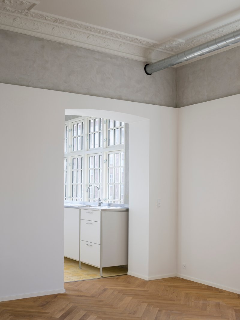 View through white-walled rooms with herringbone wood floors toward a kitchenette beneath gridded casement windows