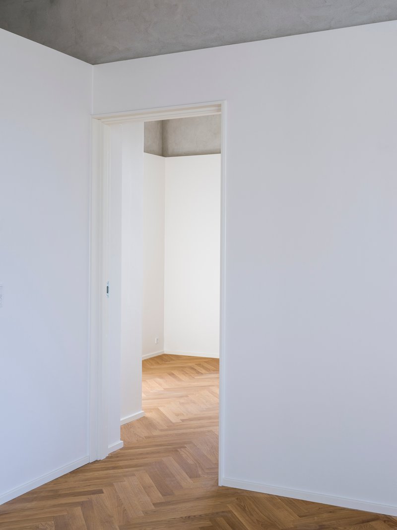Doorway framed in white trim connecting rooms with herringbone parquet and exposed concrete ceiling beam