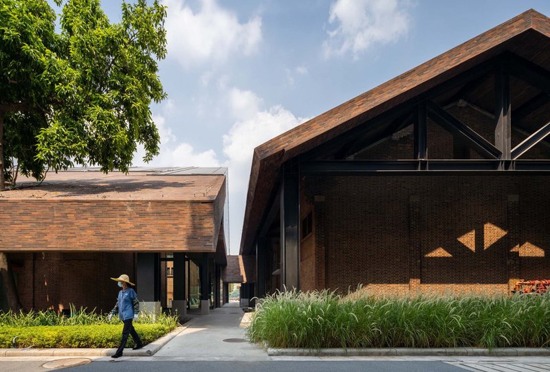 Brick volumes with timber roofs framing a paved pathway, with a person walking past planted beds