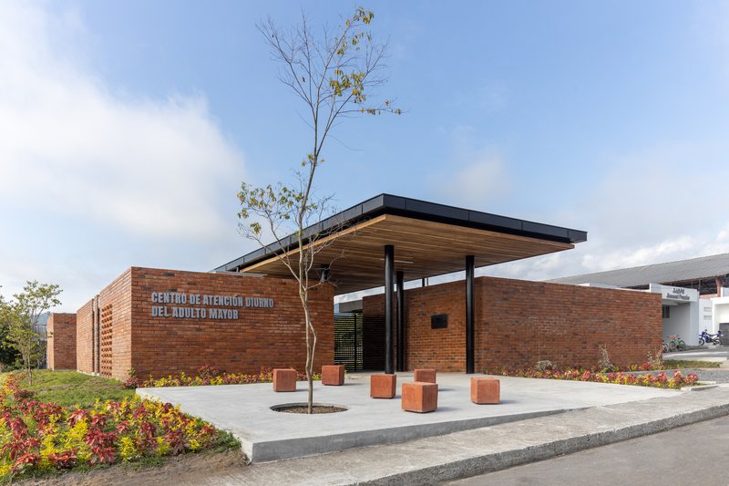 Red brick facade with cantilevered timber canopy sheltering a concrete plaza with planted tree