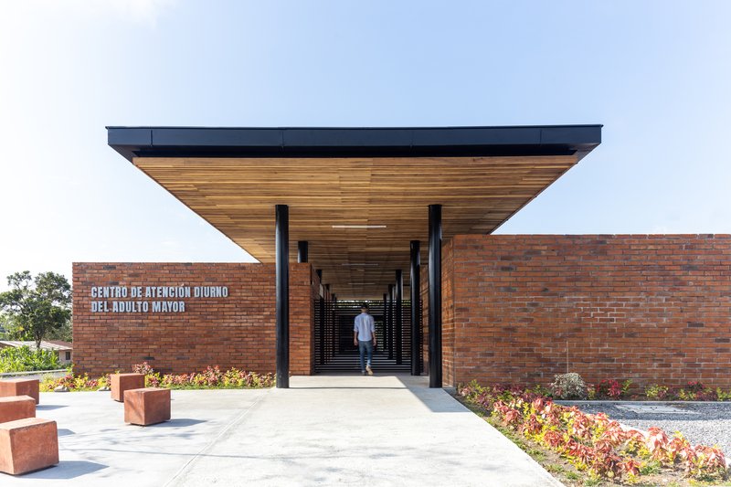 Entry corridor with timber slat ceiling and black steel columns framing a figure walking toward the courtyard