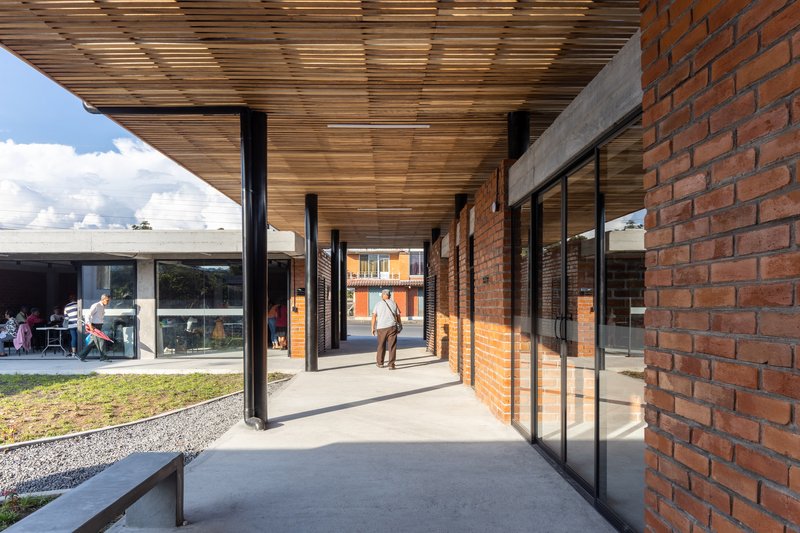 Linear covered walkway with timber ceiling, brick walls and glazed doors casting shadows under afternoon sun