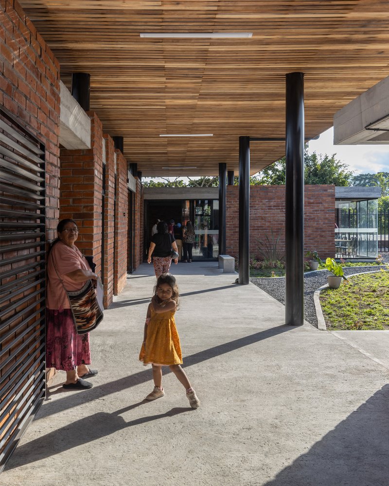 Covered walkway with slatted timber ceiling and black steel columns where a child walks in afternoon light