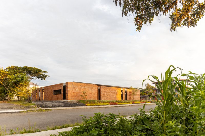 Street view of the brick volume with vertical window slots framed by trees and vegetation at golden hour