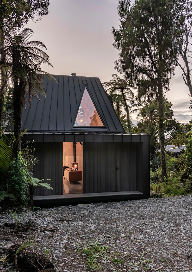 Steep-pitched black metal roof with triangular window glowing at twilight among tropical trees