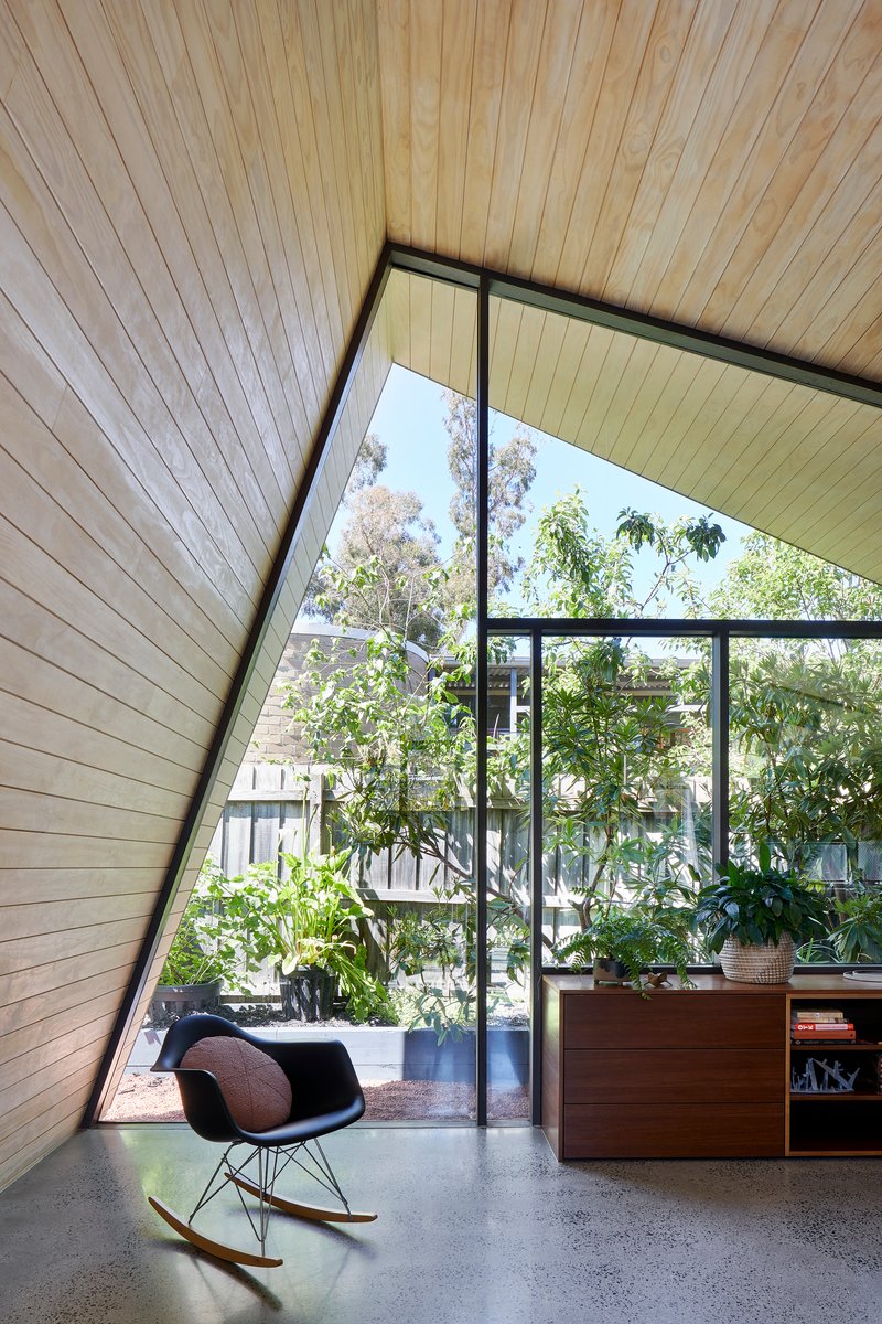 Corner room with raking timber ceiling and glazed wall overlooking a planted courtyard with rocking chair
