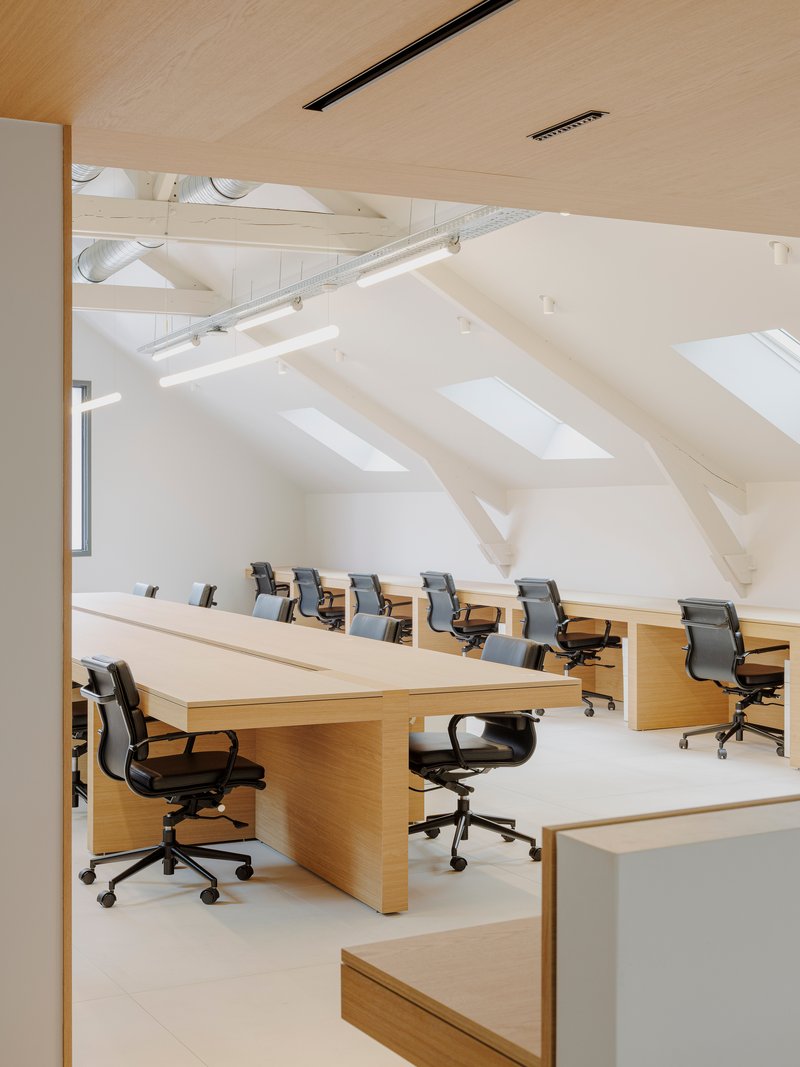 Open-plan office with rows of oak desks and office chairs under exposed white-painted roof trusses