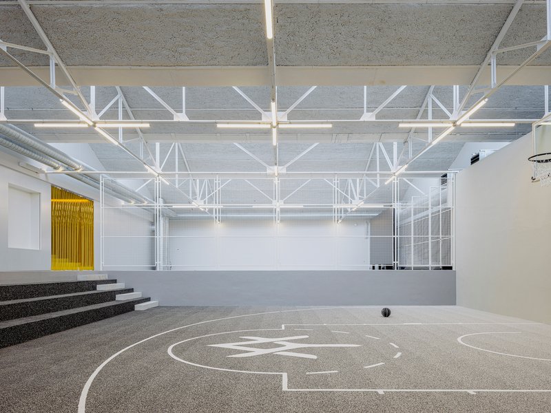 Gymnasium with white floor markings, exposed steel trusses, and tiered concrete seating along one wall