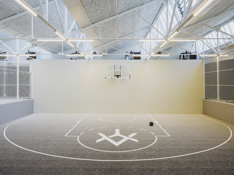 Indoor basketball court with white structural trusses overhead and tall glazed walls on both sides