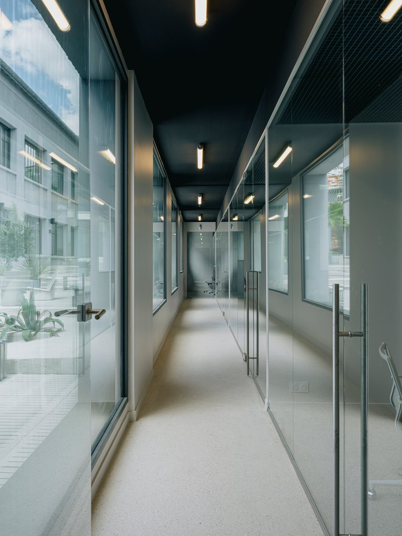 Glass-walled corridor with black ceiling and linear lighting overlooking an exterior courtyard with plantings
