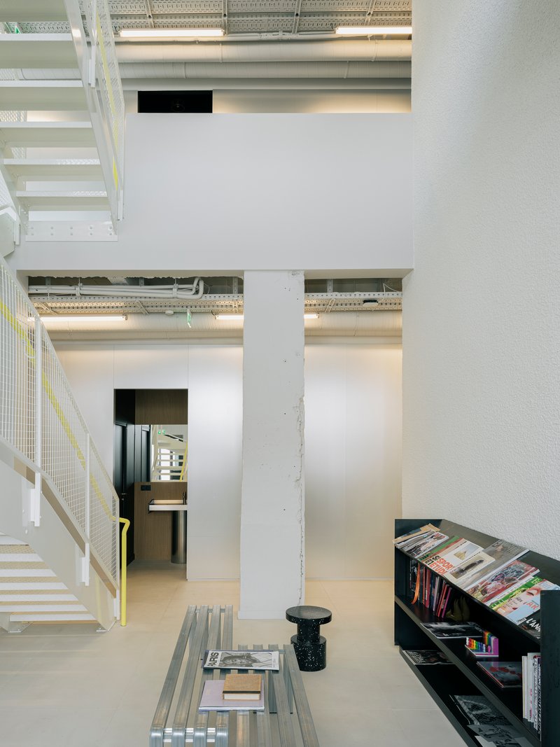 Double-height interior with exposed ductwork, concrete columns, metal stair, and magazine shelving below mezzanine