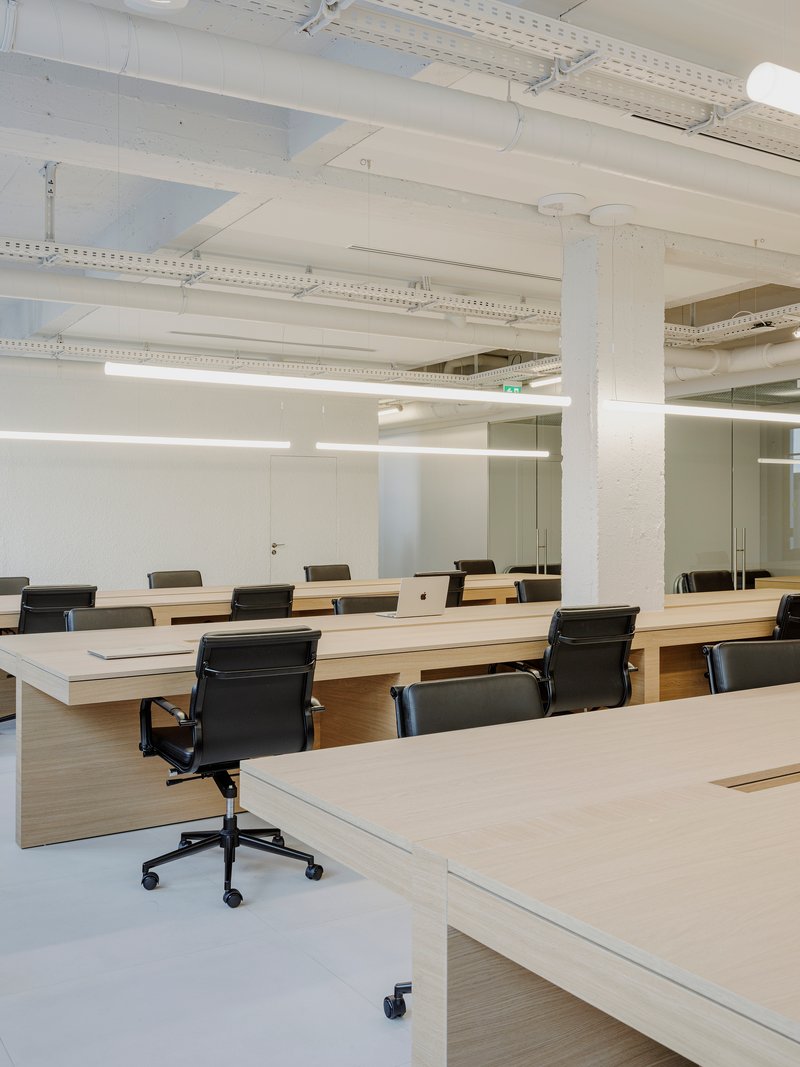 Rows of light wood desks with black office chairs under painted white structural beams and ductwork