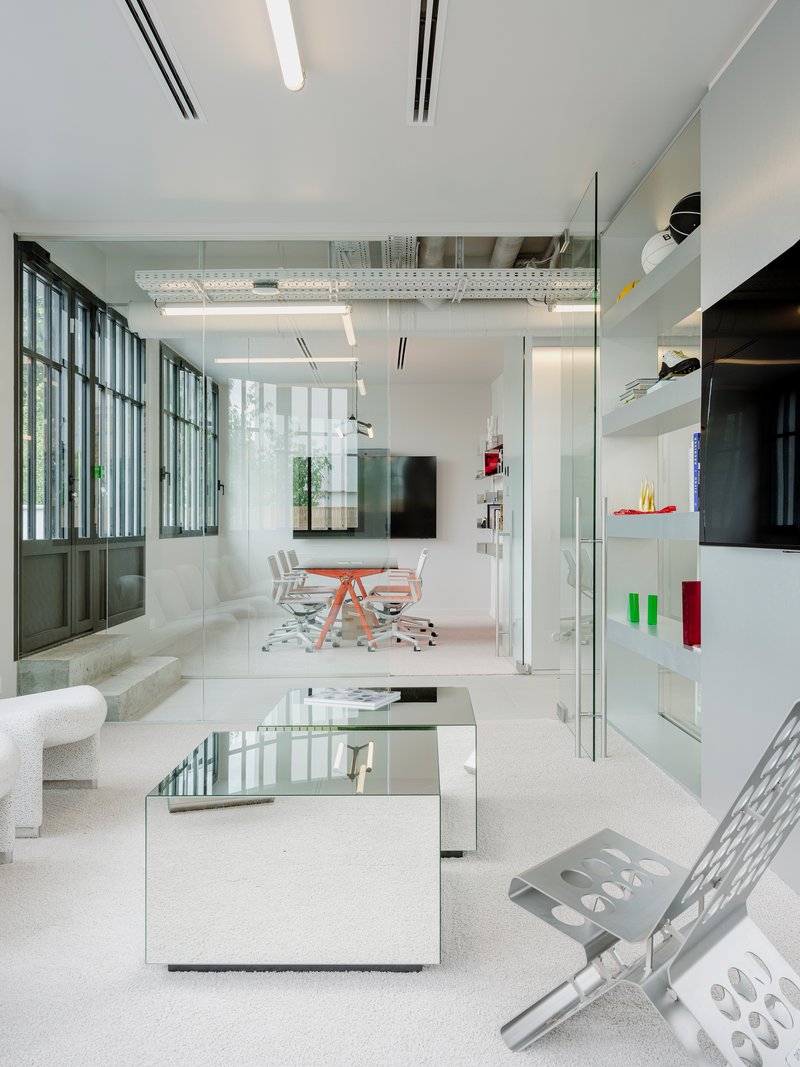 Double-height office space with glass partitions, mirrored coffee table, and steel-framed windows overlooking greenery