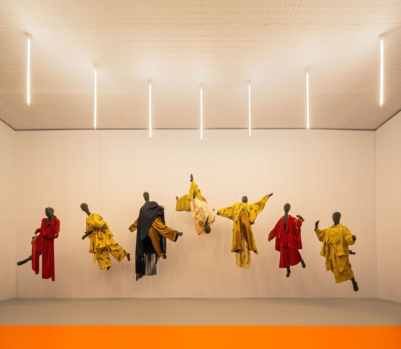 Exhibition wall with suspended garments in yellow and red under linear ceiling lights and orange floor stripe