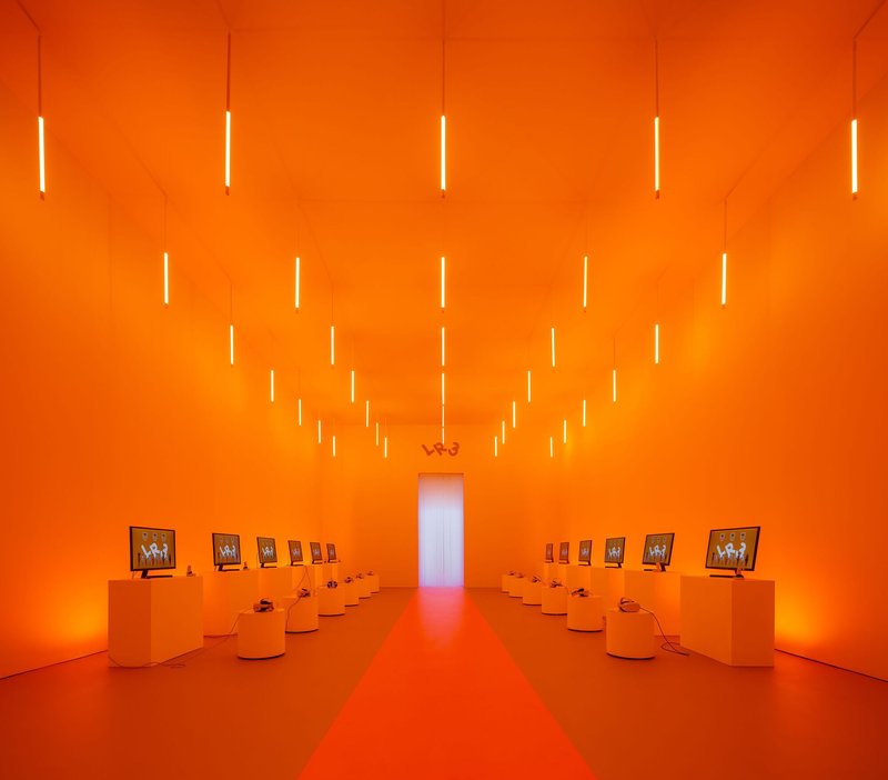 Gallery space with orange walls, vertical light tubes suspended from ceiling, and low display plinths