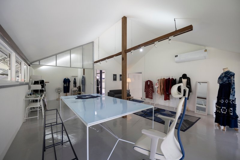 Interior workspace with white worktable, exposed timber beams crossing overhead, and garments displayed on wall