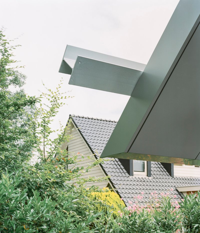Cantilevered metal roof planes extending over neighboring tiled roof and garden plantings