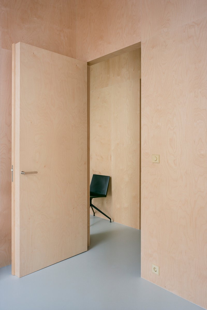 Pale plywood-lined hallway with flush door and glimpse of black chair beyond