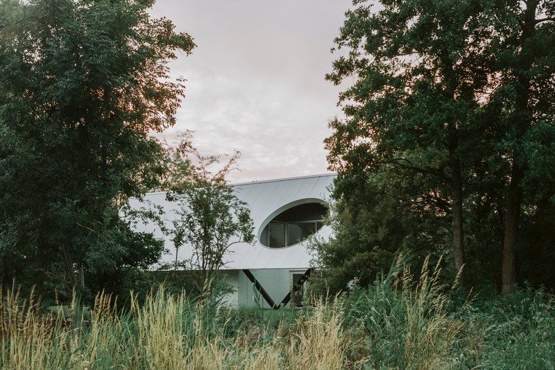 White corrugated metal facade with circular opening framed by tall trees and wild grasses at dusk