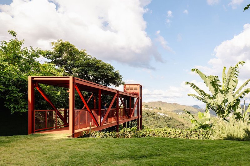 Red steel structural frame with diagonal bracing extending over a grass lawn with banana plants and distant hills