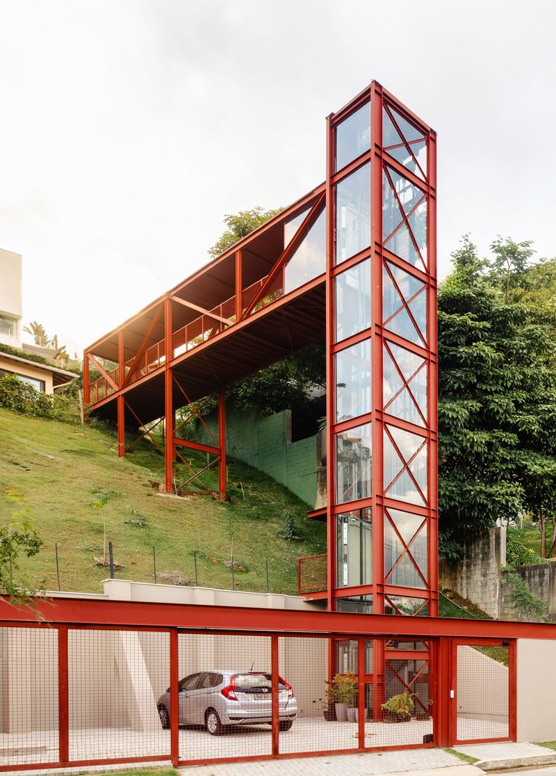 Vertical glass circulation tower enclosed in red steel bracing above a carport with wire mesh gates