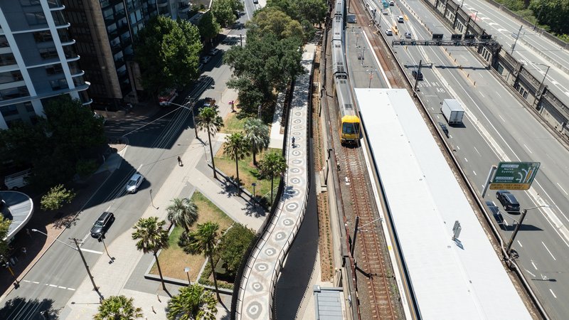 Overhead view of the elevated railway platform with white canopy roof running between landscaped pathways and highway