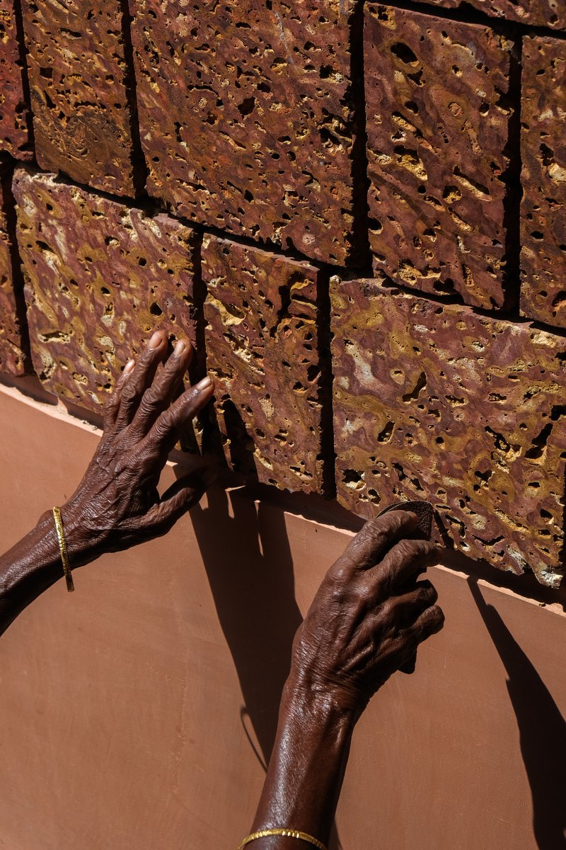 Close-up of hands touching textured laterite brick wall showing irregular surface and deep mortar joints in sunlight