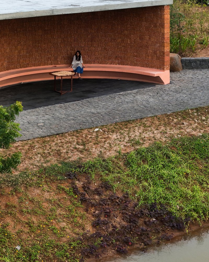 Curved terracotta bench against laterite brick wall with seated figure on dark stone paving beside pond
