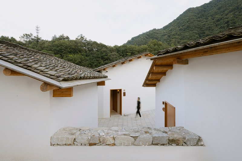 Courtyard passage with white plastered walls, timber eaves, and fieldstone outcrop with a figure passing through