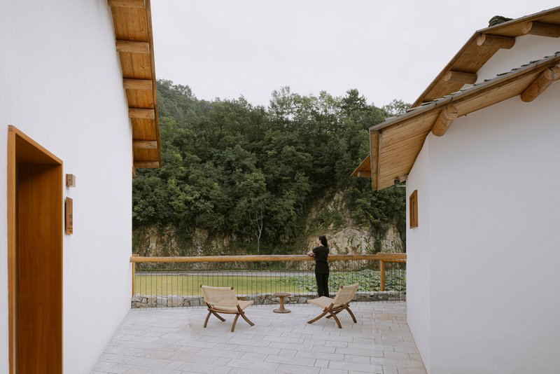 Courtyard terrace with wooden lounge chairs facing a pond and forested hillside under overcast skies