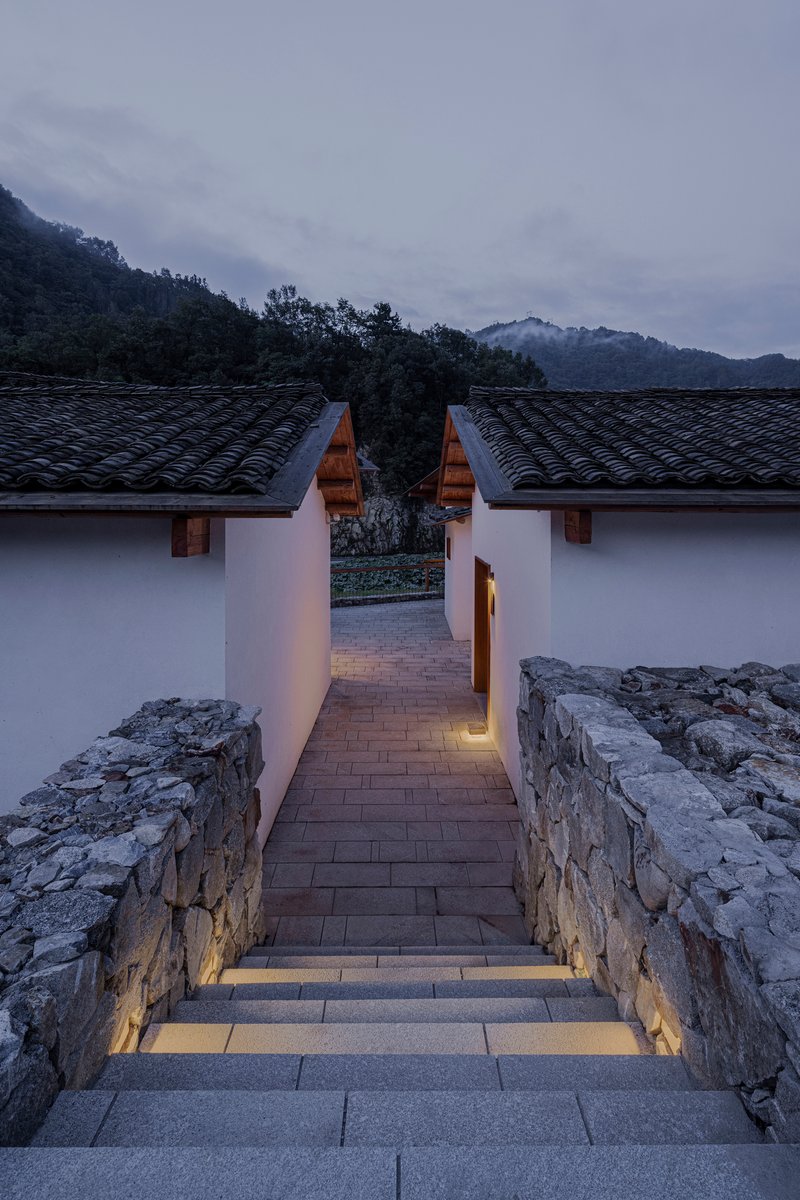Stone staircase with recessed lighting descending between white walls toward tiled roofs at dusk