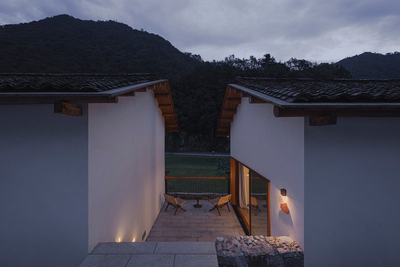 Entry courtyard between two white volumes with timber eaves and uplighting against mountain backdrop at twilight