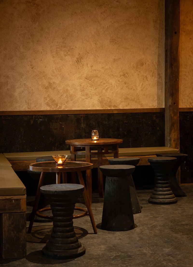 Low timber tables and turned stools against a two-tone plaster wall lit by candlelight