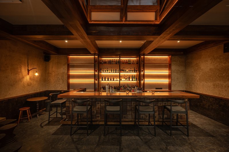 Bar interior with timber-beamed coffered ceiling, skylights, backlit bottle shelves and stools around a counter