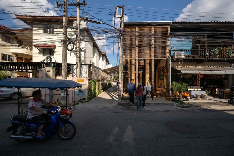 Street view of a metal screen facade with pedestrians and a motorcyclist passing beneath overhead wires