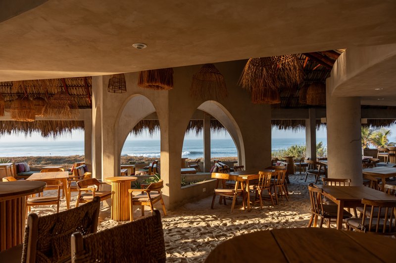 Dining area with earthen arches framing ocean views and dappled sunlight across sand floor