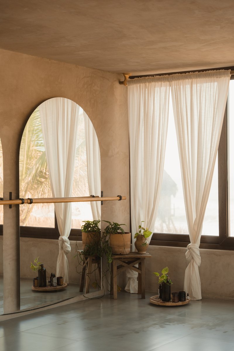 Arched mirror with timber shelf and potted plants against plaster wall with sheer curtains filtering daylight