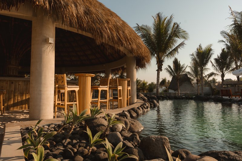 Poolside bar pavilion with thatched roof, concrete columns and timber stools beside a rock-edged lagoon at dusk