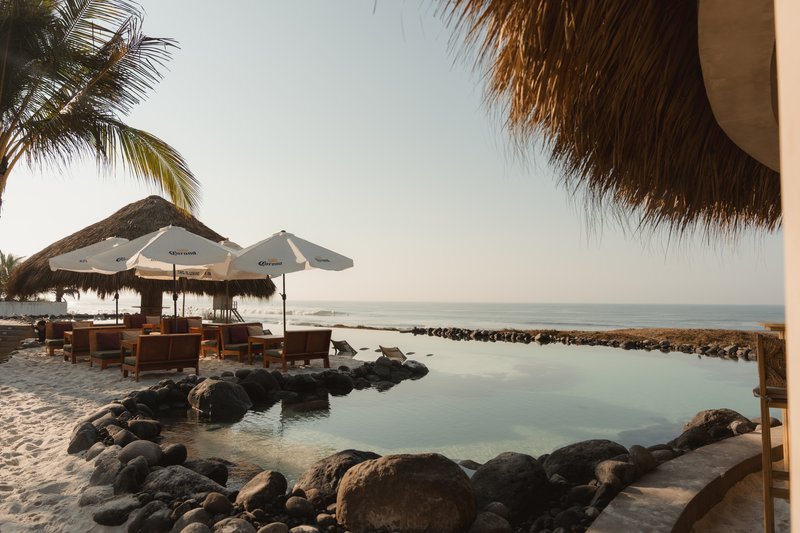 Rock-edged saltwater pool with thatch-roofed pavilion and lounge seating near the ocean at dusk