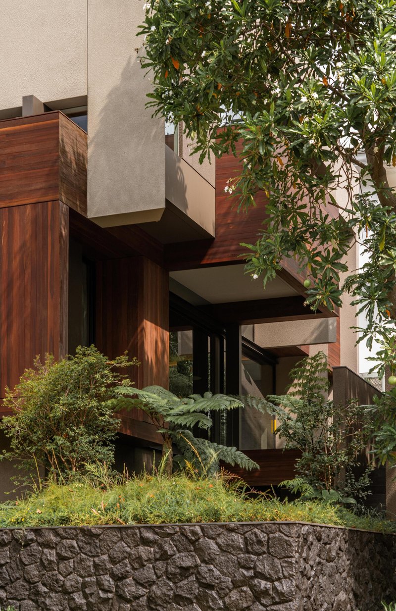Street view of stacked timber-clad volumes with planted terraces framed by trees and stone retaining wall