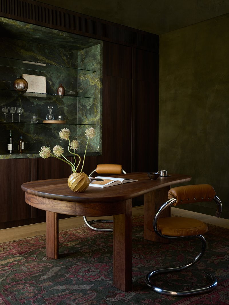 Walnut desk and cantilever chair in an olive-green room with glass shelving and dried floral arrangement