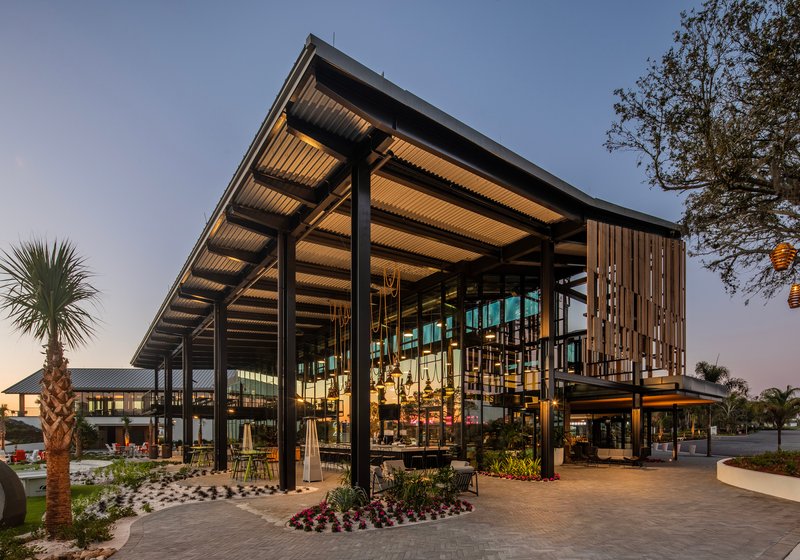 Covered pavilion with steel columns and slatted ceiling near planted palm trees at twilight