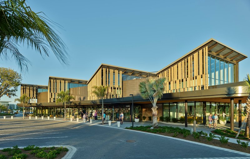 Stepped facade with vertical timber screening and clerestory glazing along a palm-lined arrival plaza