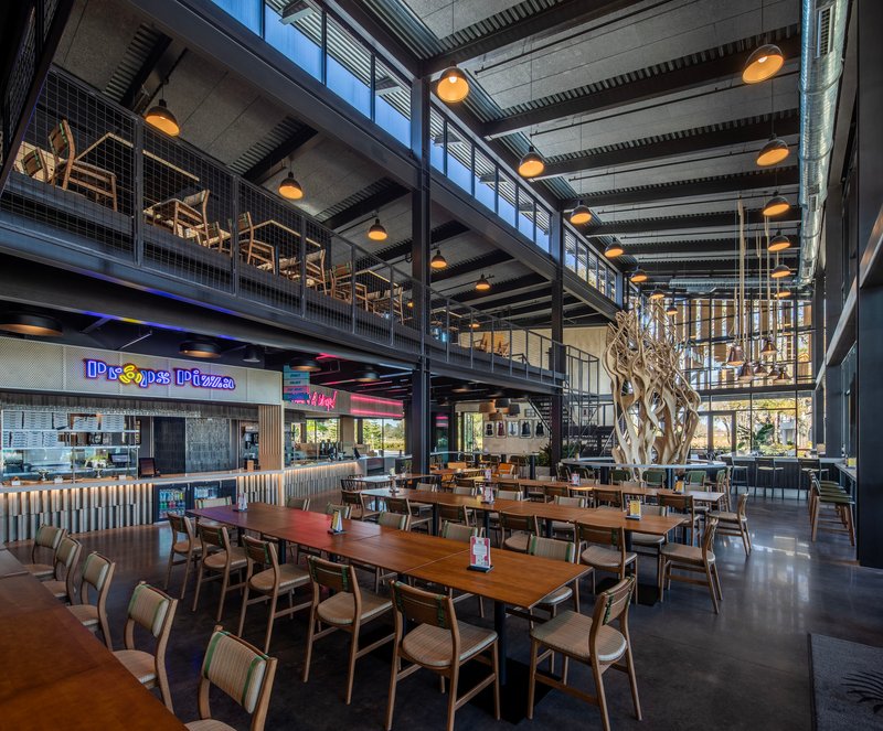 Two-story food hall with exposed concrete ceiling, steel mezzanine and a sculptural tree at center