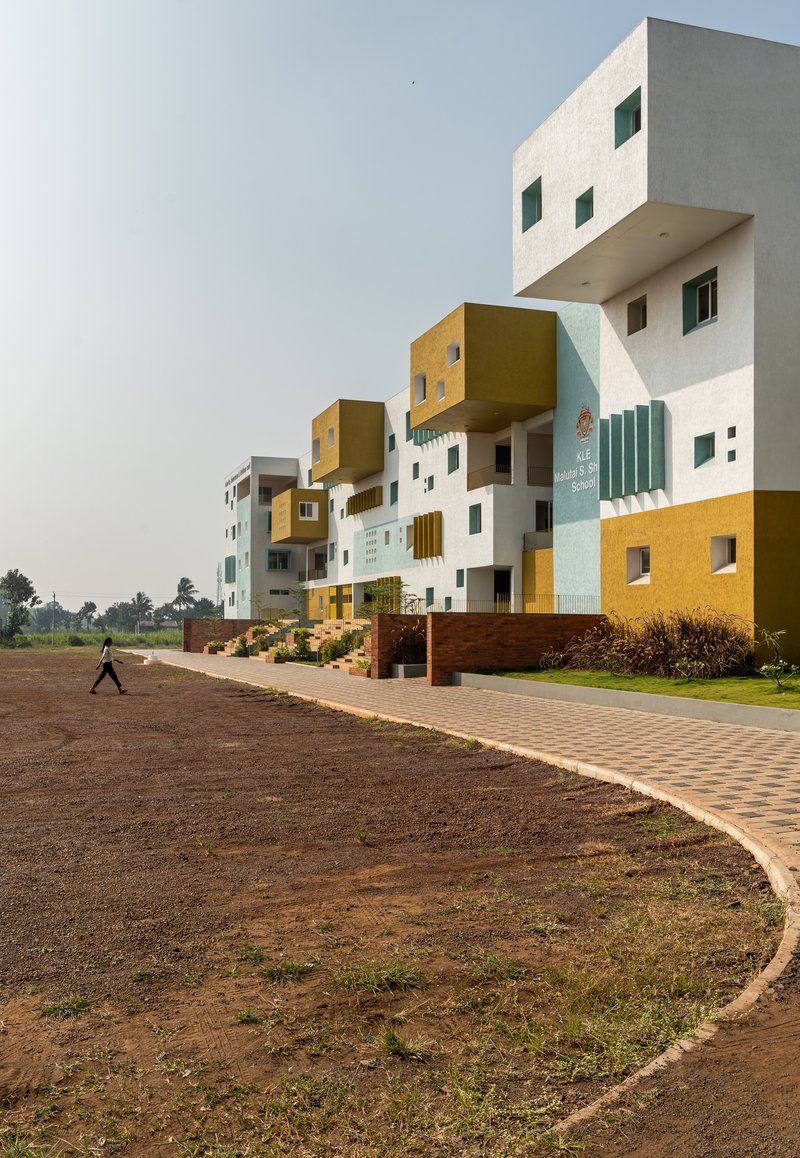 Stacked volumes clad in white, yellow and pale blue with cantilevered boxes and a person crossing the brick paved forecourt