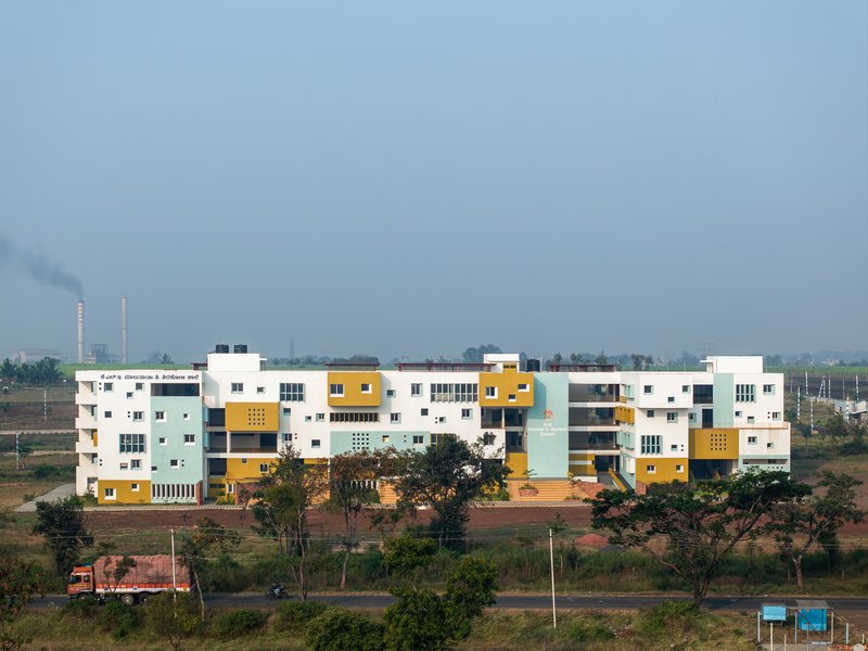 Horizontal facade of interlocking volumes in white, yellow and pale blue under a hazy sky with distant smokestacks