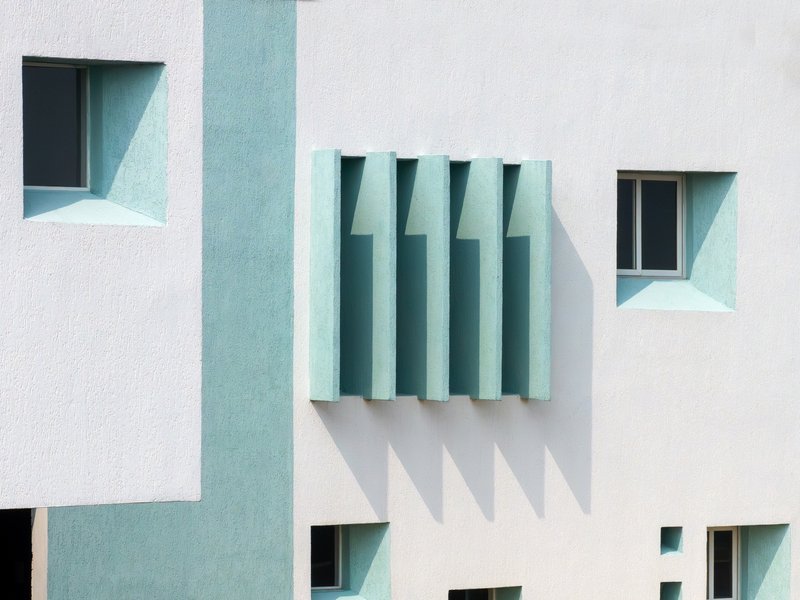 Close-up of white stucco facade with pale blue vertical louvers and recessed windows with angled reveals