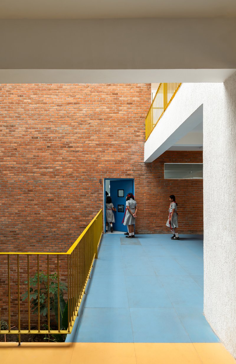Corridor with exposed brick wall, blue tile floor, and yellow metal railing framing a planted courtyard