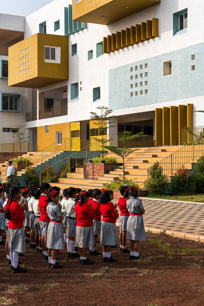 Courtyard view of white and mustard-colored volumes with children in red uniforms gathered on the paved ground